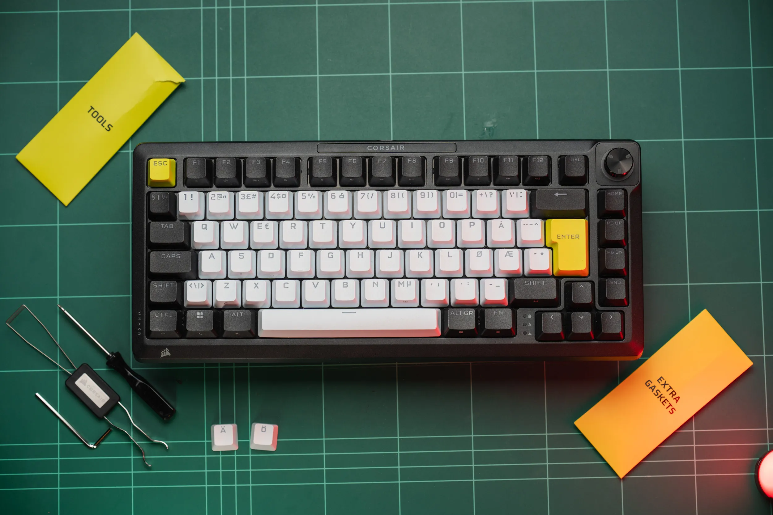 The Corsair MAKR PRO 75 on a green cutting mat with its bundled tools, the labelled tools and extra-gaskets envelopes, and the spare Nordic keycaps