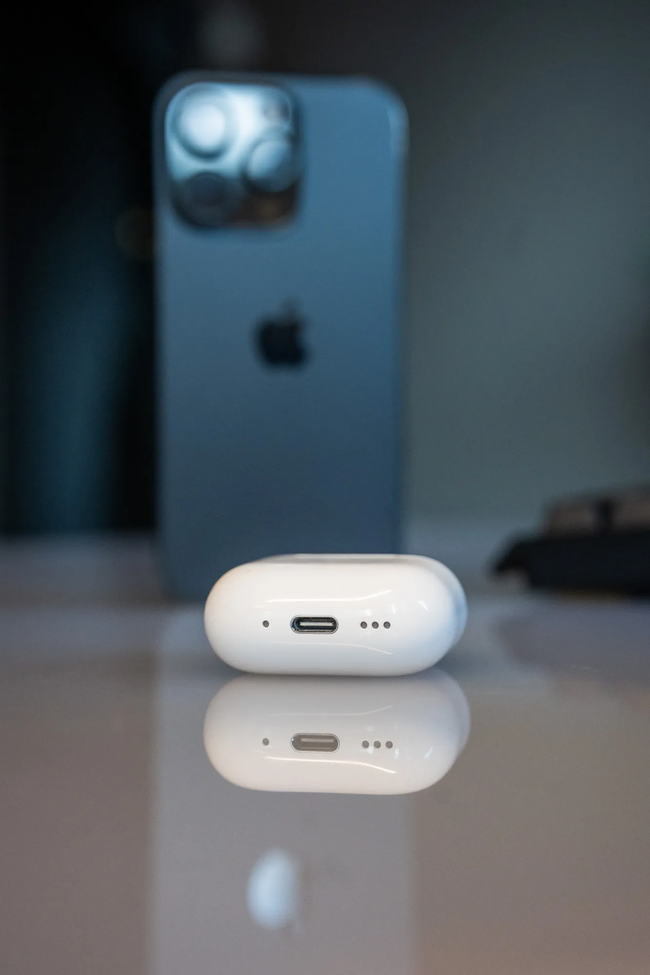 The underside of the AirPods 5 case with the USB-C port and speaker holes, an iPhone blurred in the background