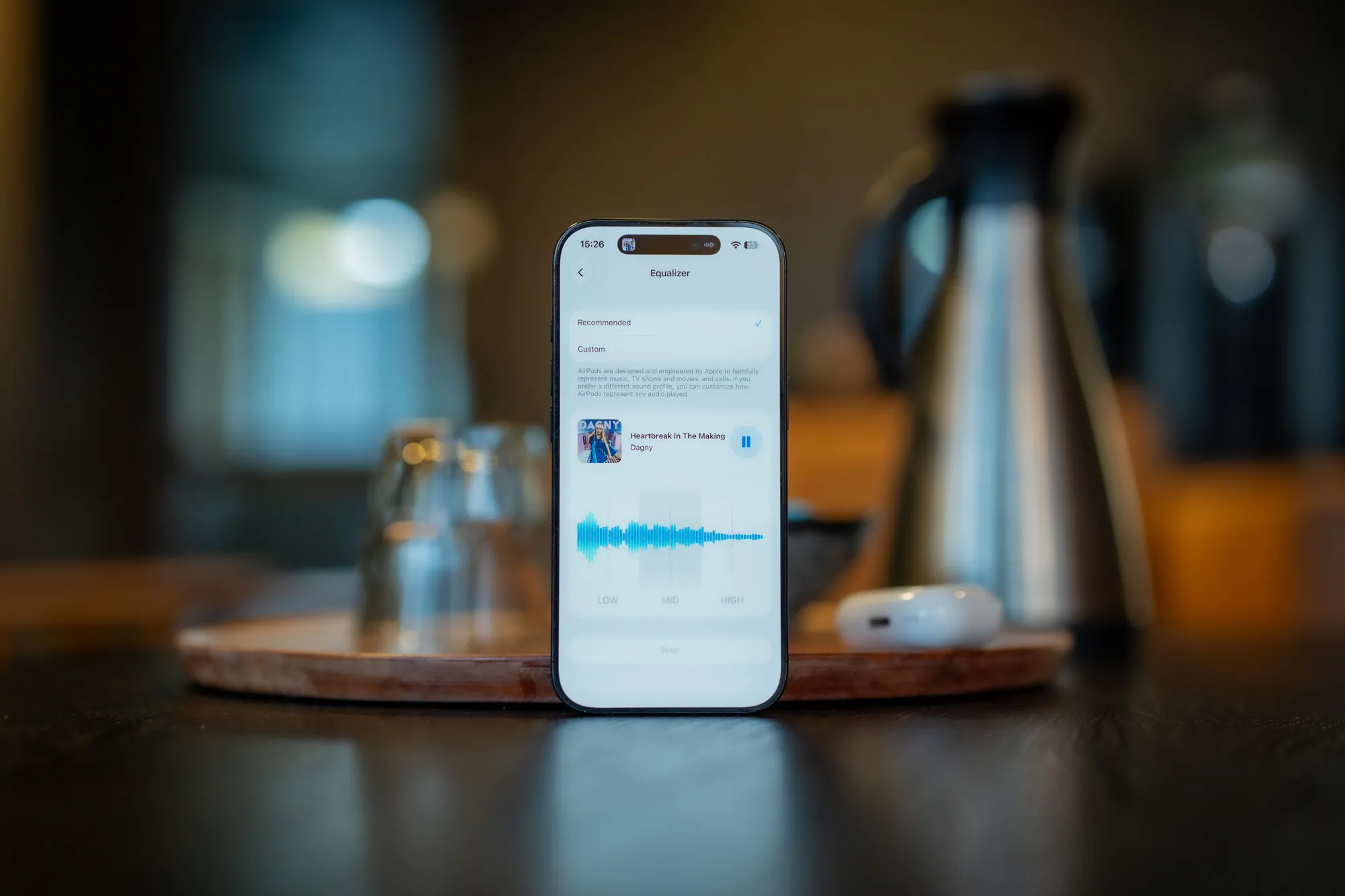 iPhone standing on a wooden board with the AirPods 5 equalizer on screen, the AirPods case and a thermos in the background