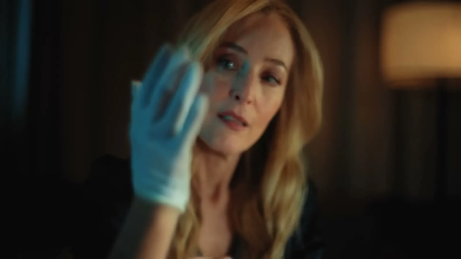 Gillian Anderson pulling on a blue latex glove in a dim hotel room