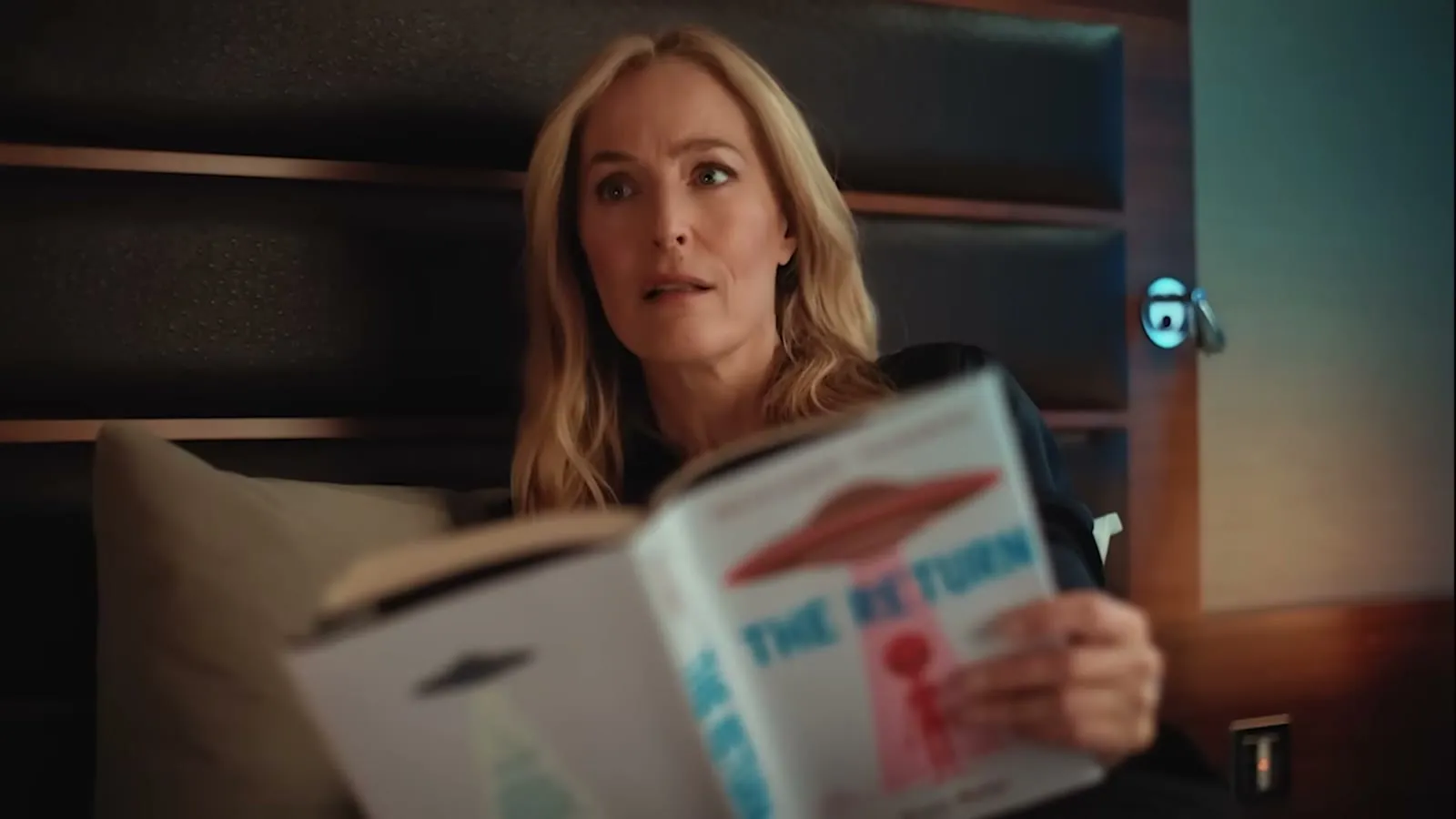 Gillian Anderson in a hotel bed reading a magazine with a UFO on the cover